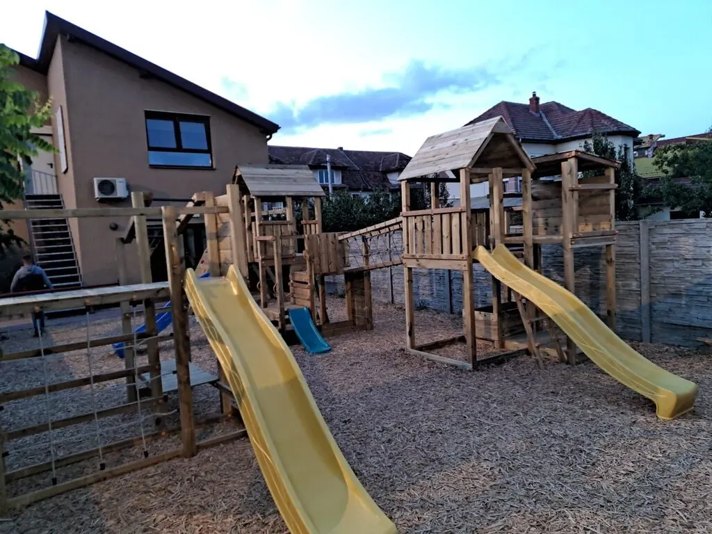 Zona de joacă din curtea grădiniței, cu două tobogane galbene și turnuri de lemn cu acoperiș