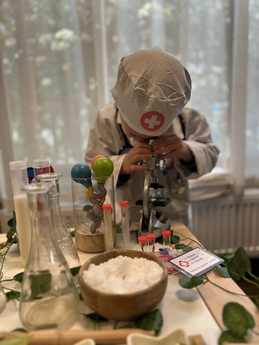 Copil costumat în doctor privește printr-un microscop, la o masă cu eprubete și plante